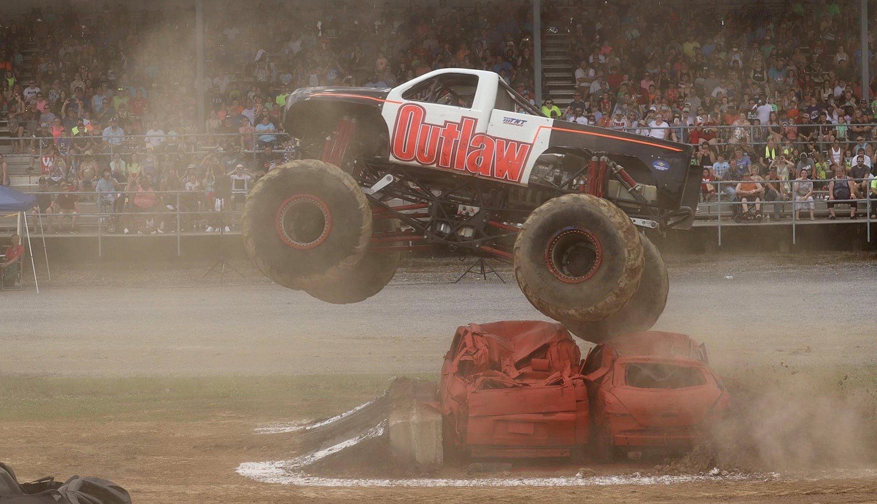 Monster truck jumping over crushed cars, raising dust at an outdoor event.