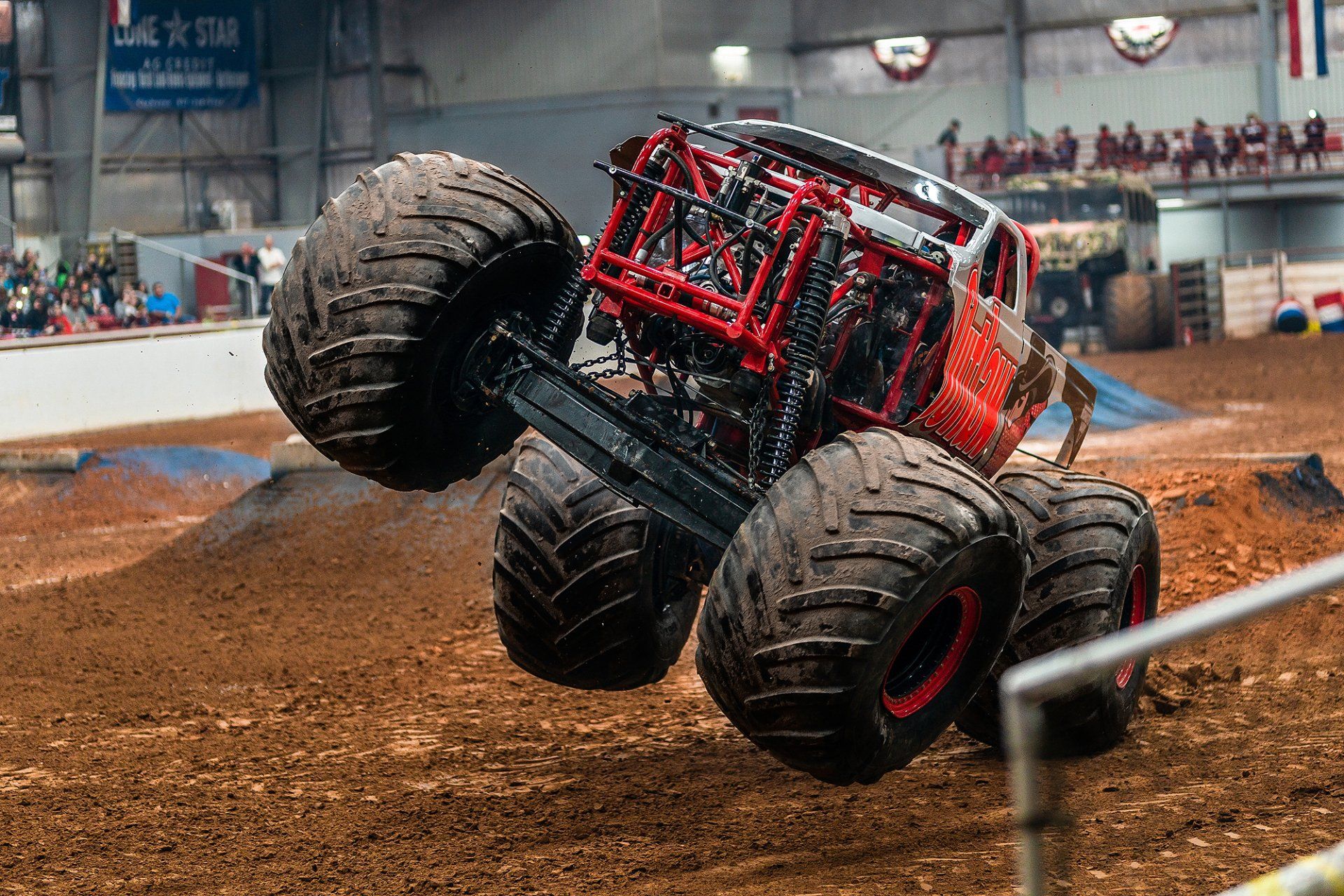 Monster truck in mid-air, red and black chassis, large tires, brown dirt track, indoor arena with spectators.