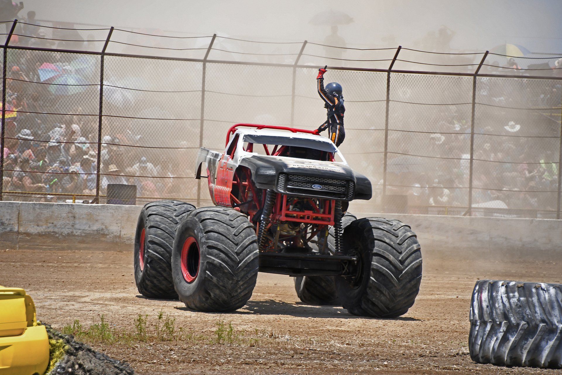 Monster truck with a driver on the roof, waving to the audience. Red and black truck on dirt track.