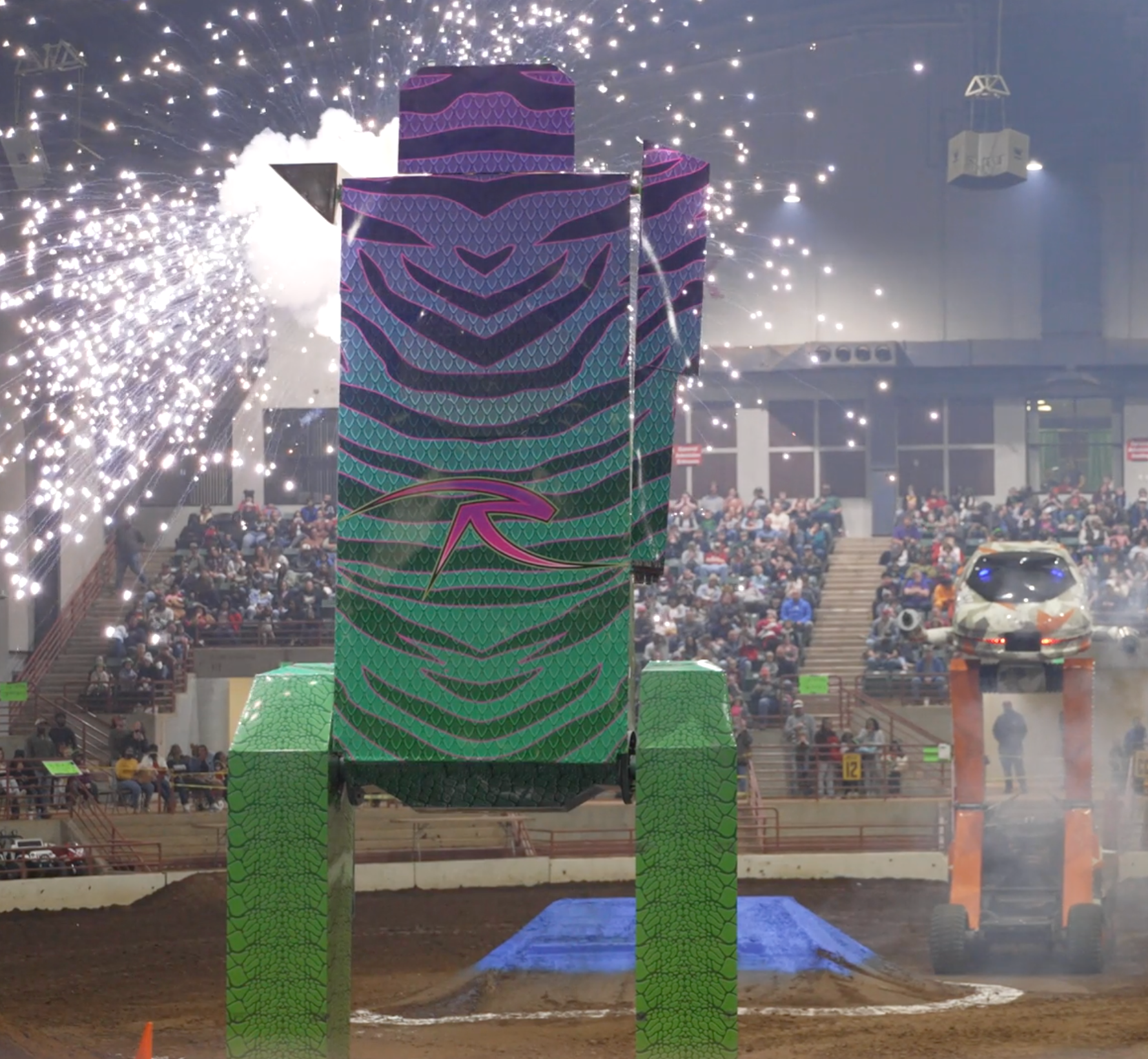 Monster truck show: A large, zebra-striped, green and purple robot prop as a car jumps over a ramp. Fireworks in the background.