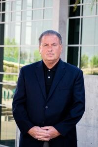 A man in a suit and black shirt is standing in front of a building with his hands folded.