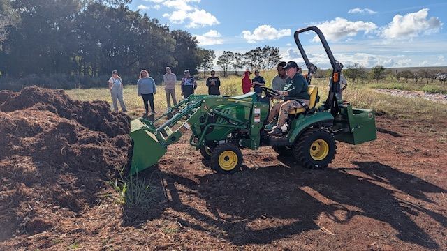 Green tractor scooping mulch in a field, with people standing nearby under a partly cloudy sky.