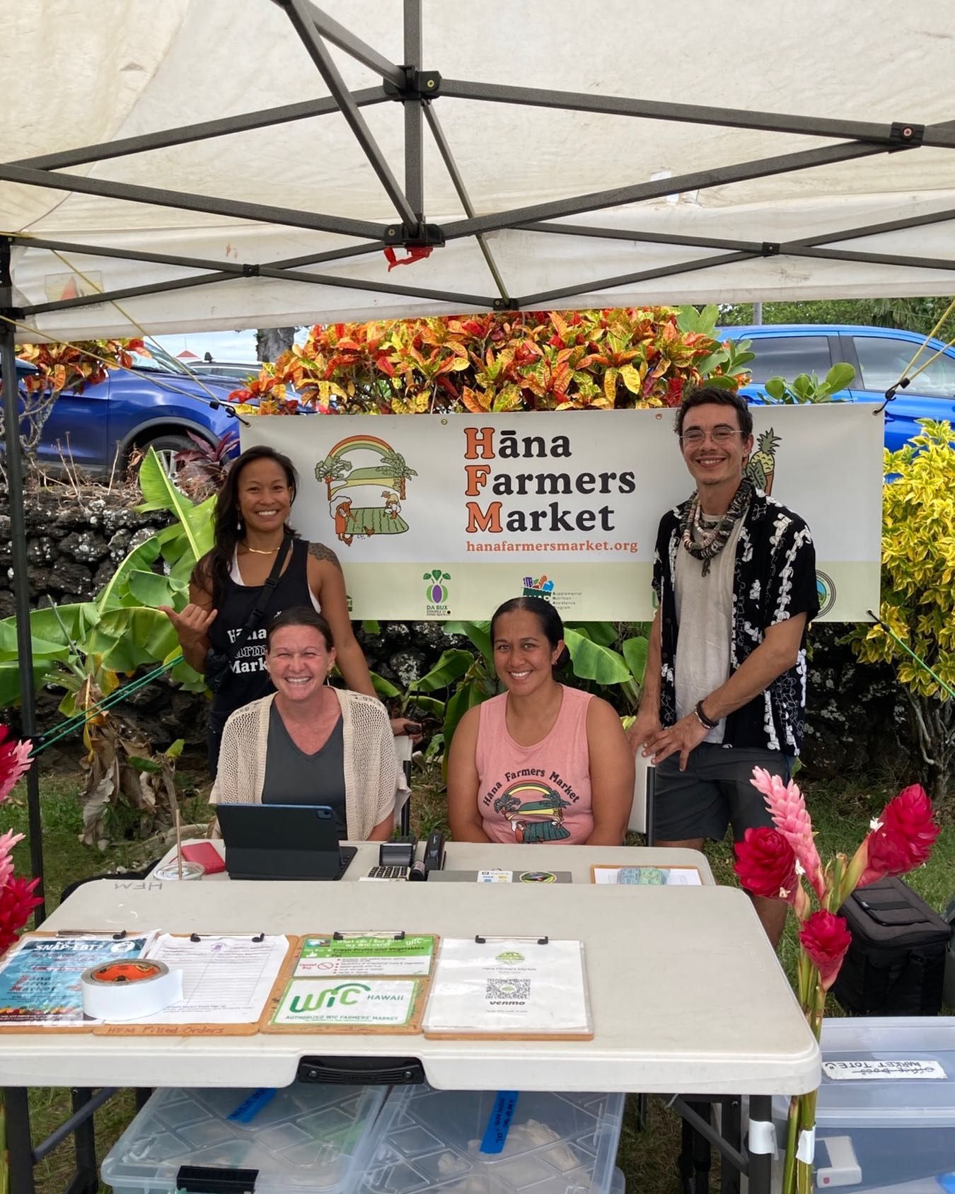 Farmers market booth under a white canopy with four people behind a table and colorful signs.