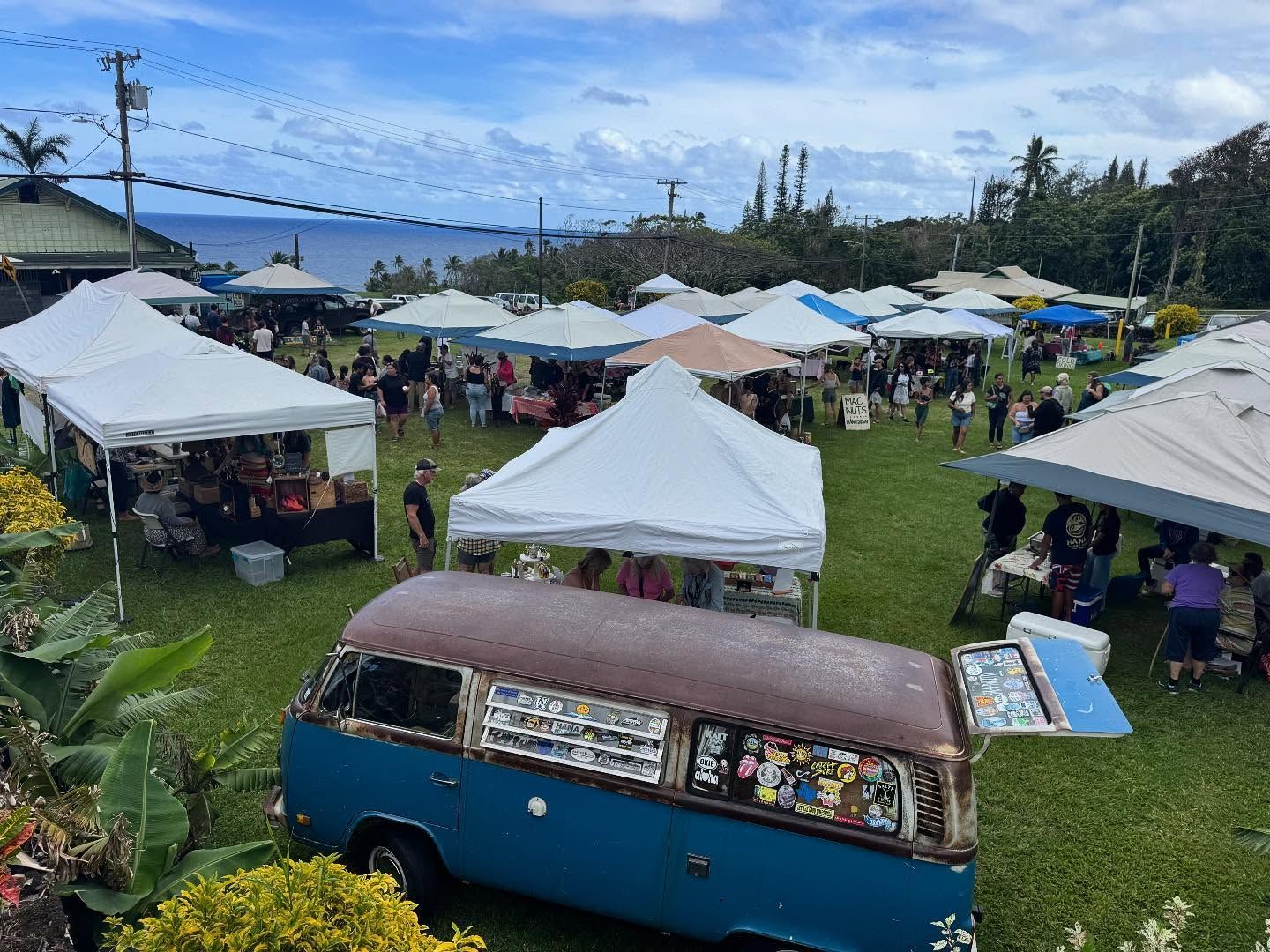 Outdoor market with white tents on a grassy field, viewed from a blue van overlooking a waterfront.
