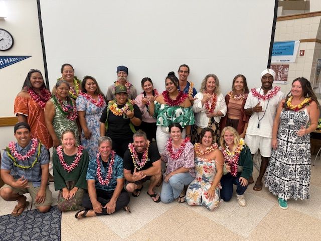 Group of people posing indoors, many wearing colorful leis, in front of a projection screen.