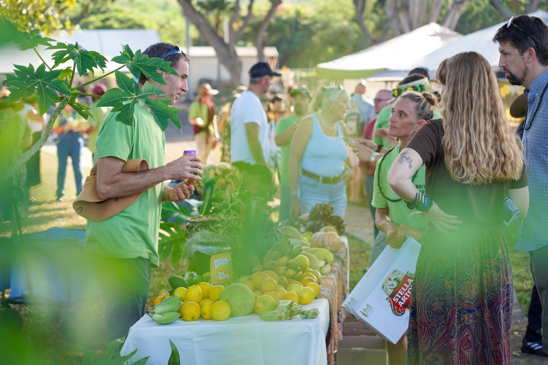 People browsing an outdoor market stall with fresh produce and flowers in bright sunlight