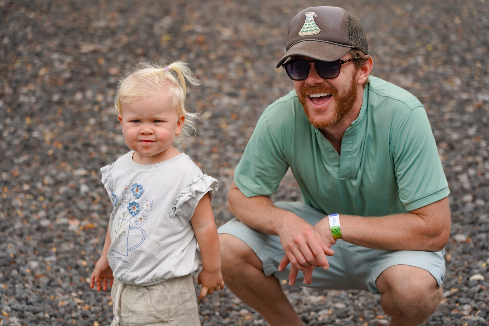 Two people crouch on a rocky beach, smiling in casual outdoor clothes and sunglasses.