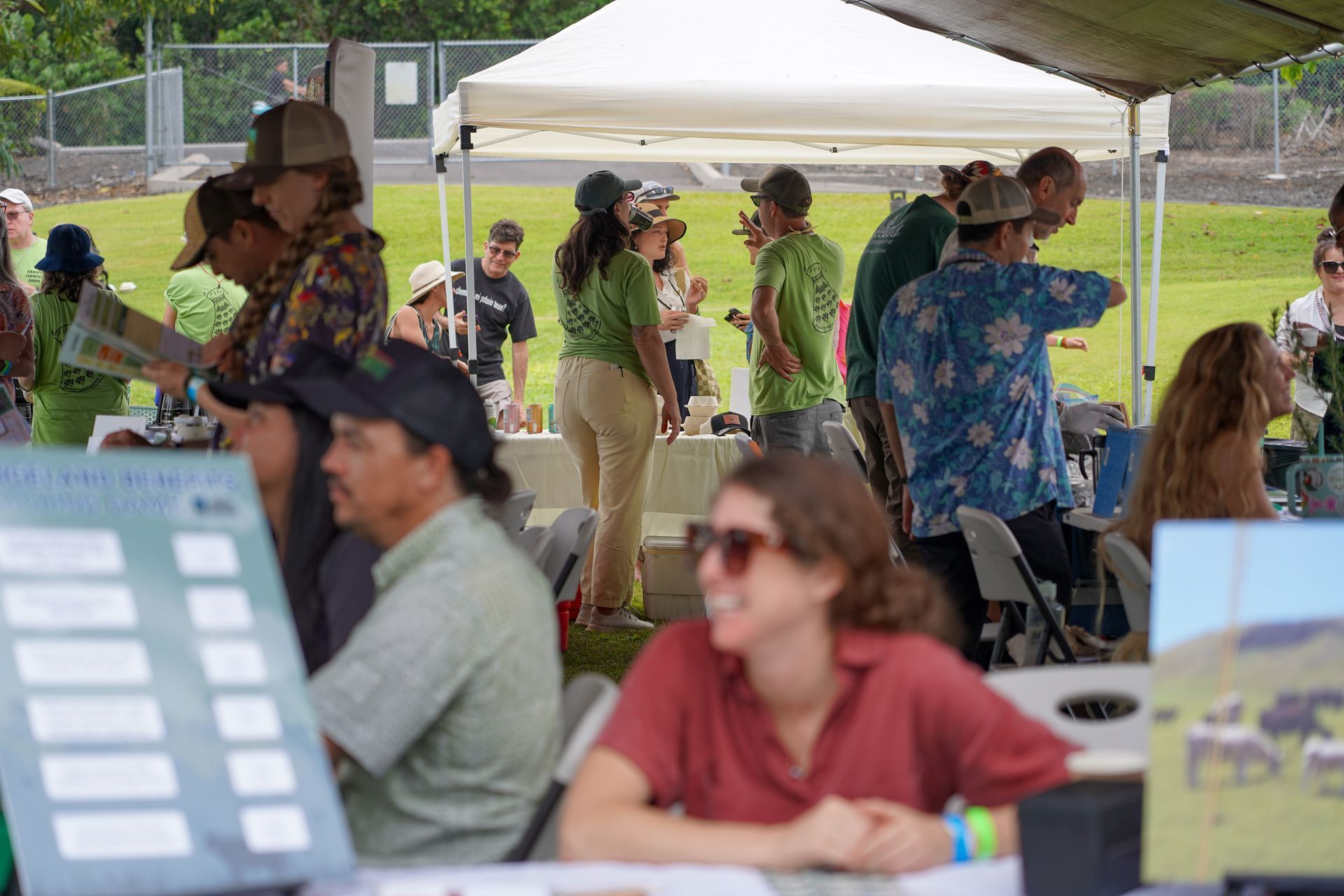 People gathered under a white tent at an outdoor event, talking and sitting at tables.