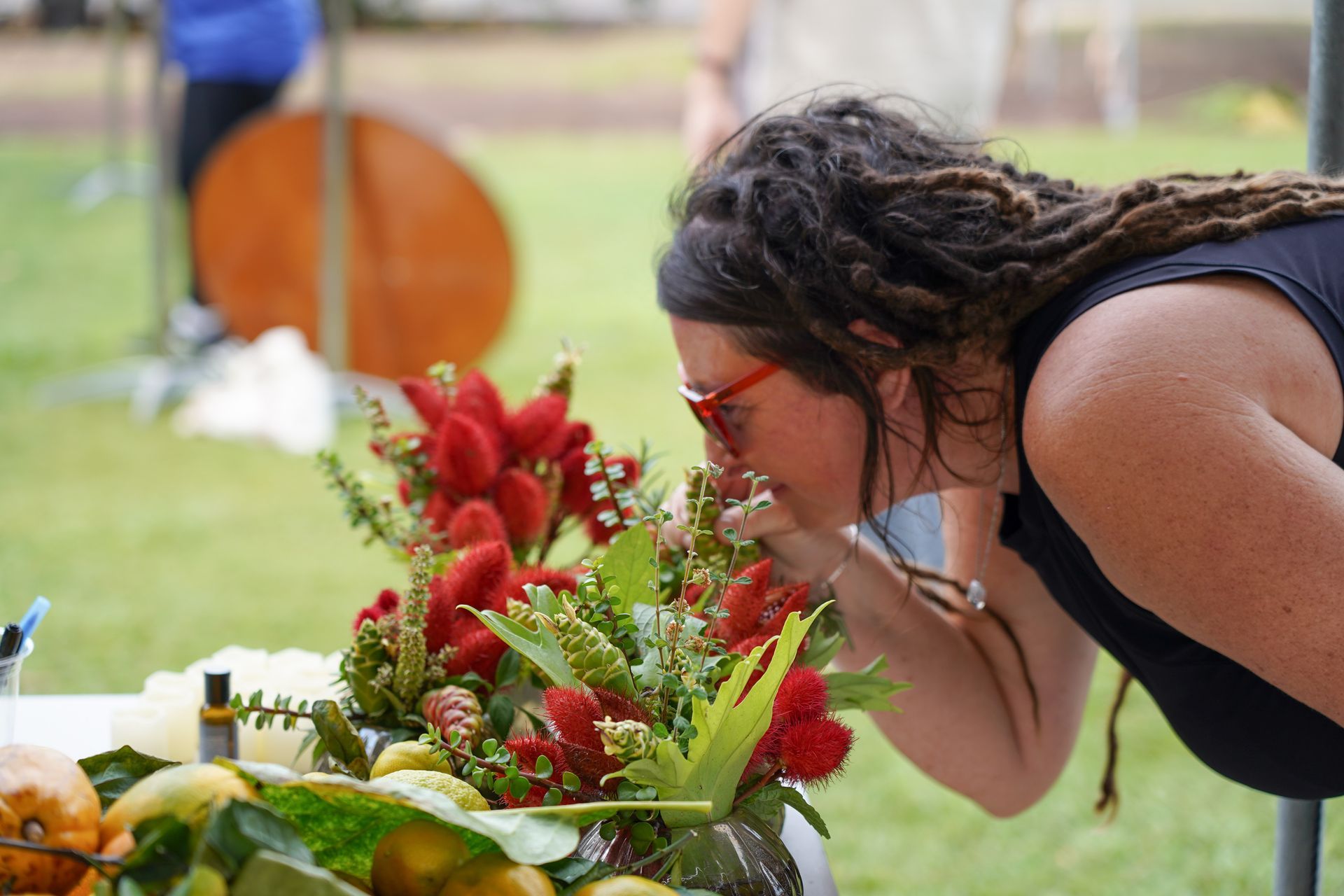Woman in black leans over a red flower arrangement outdoors, smelling the flowers at a memorial.