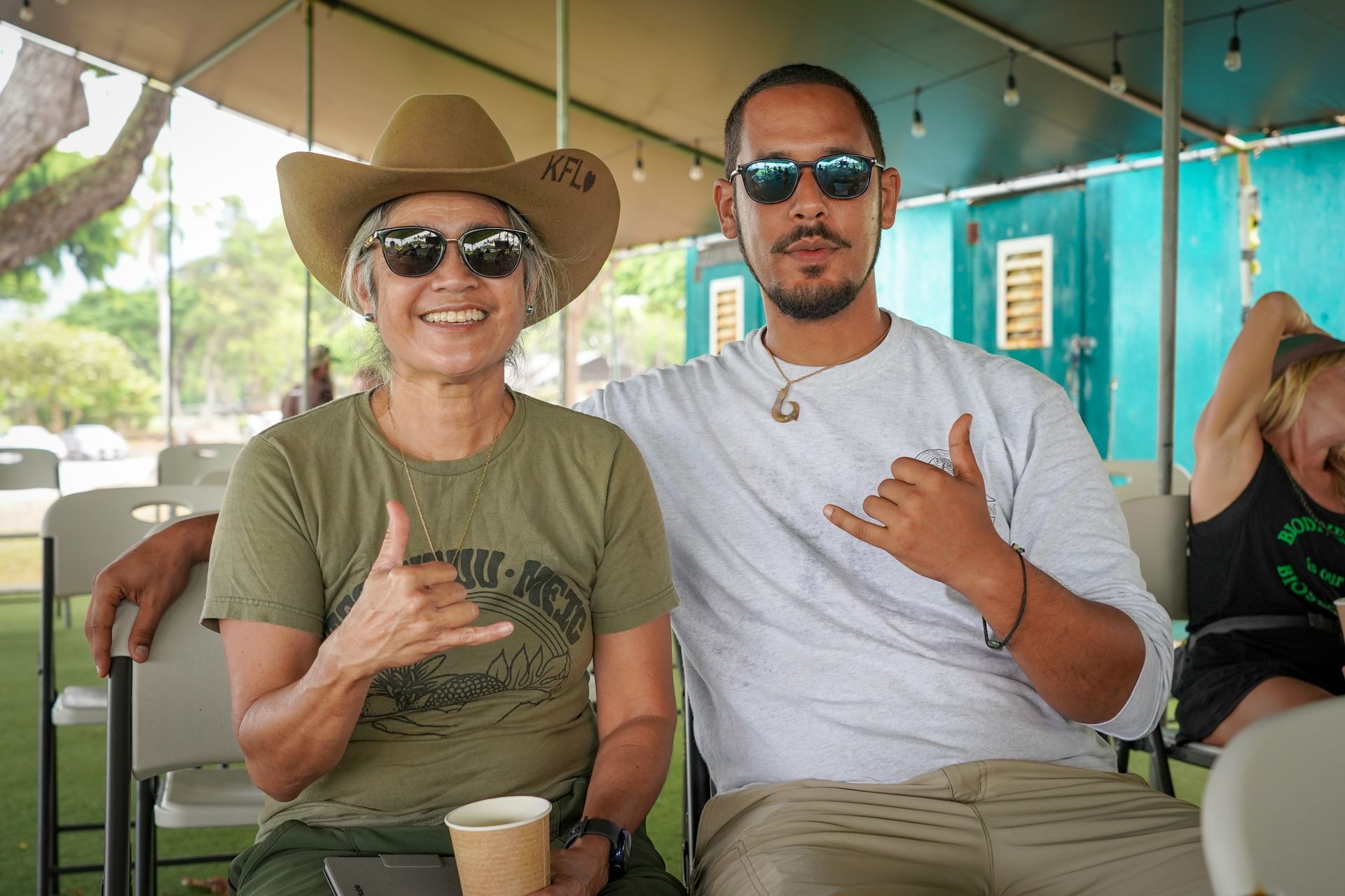Two people sitting under a tent, smiling and making shaka hand gestures, one in a hat and sunglasses, one in sunglasses.