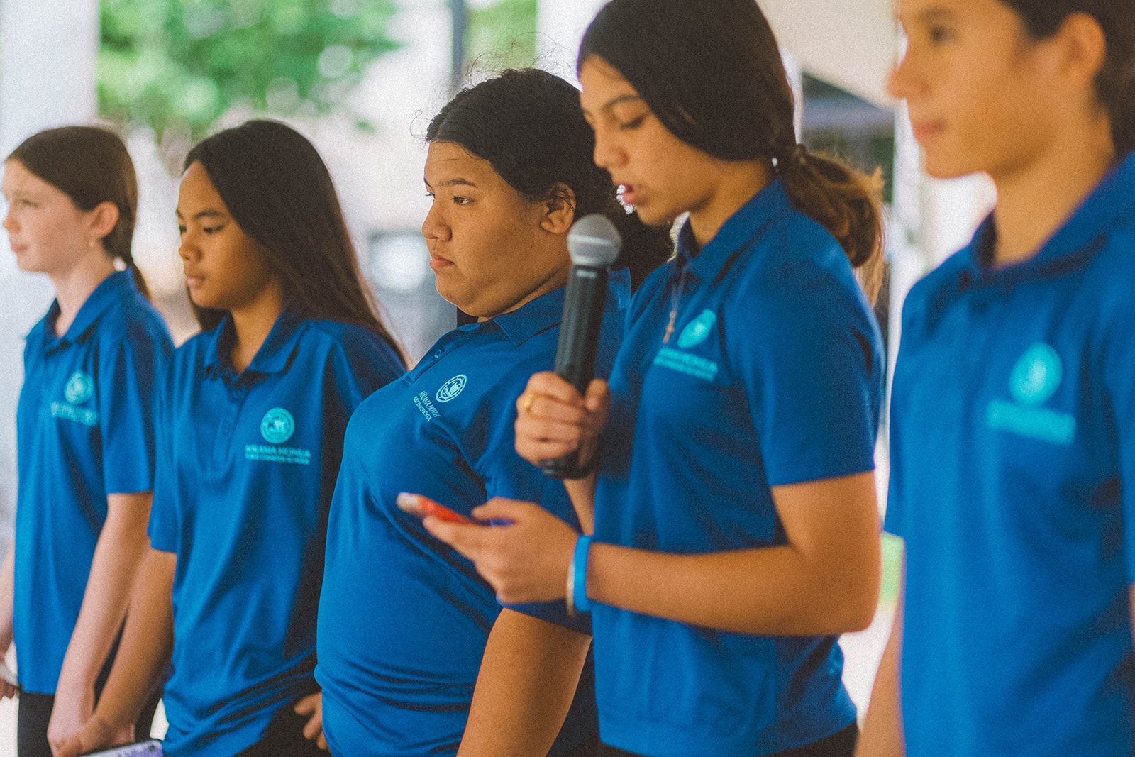 Students in blue shirts standing in a row, one speaking into a microphone