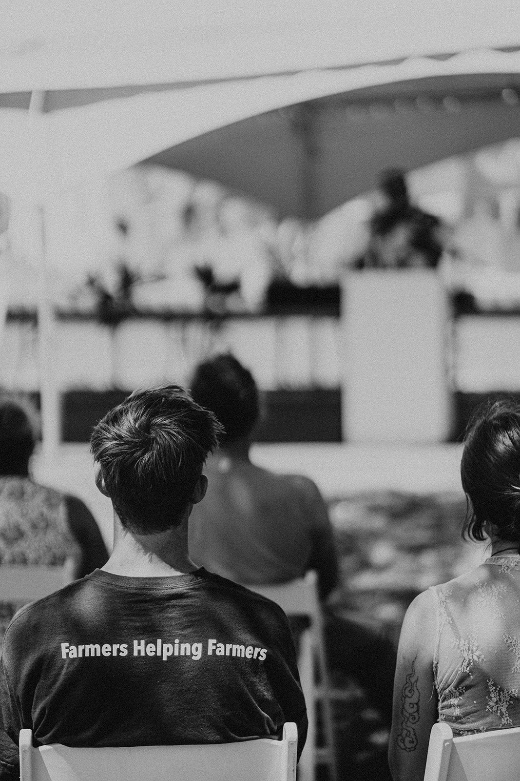 Crowd seated outdoors facing a stage, with a black shirt reading “Farmers Helping Farmers.”