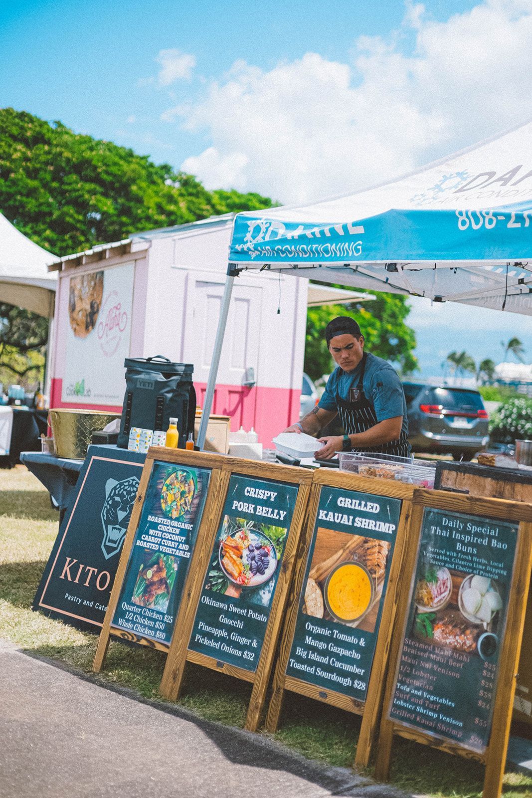 Outdoor food stall with a vendor behind menu boards under a blue canopy, surrounded by greenery.