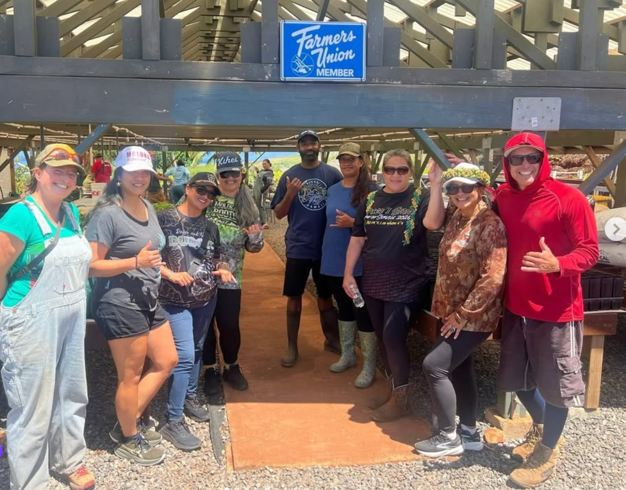 Group of people posing under a wooden shelter at a sunny outdoor event, with a blue sign overhead.
