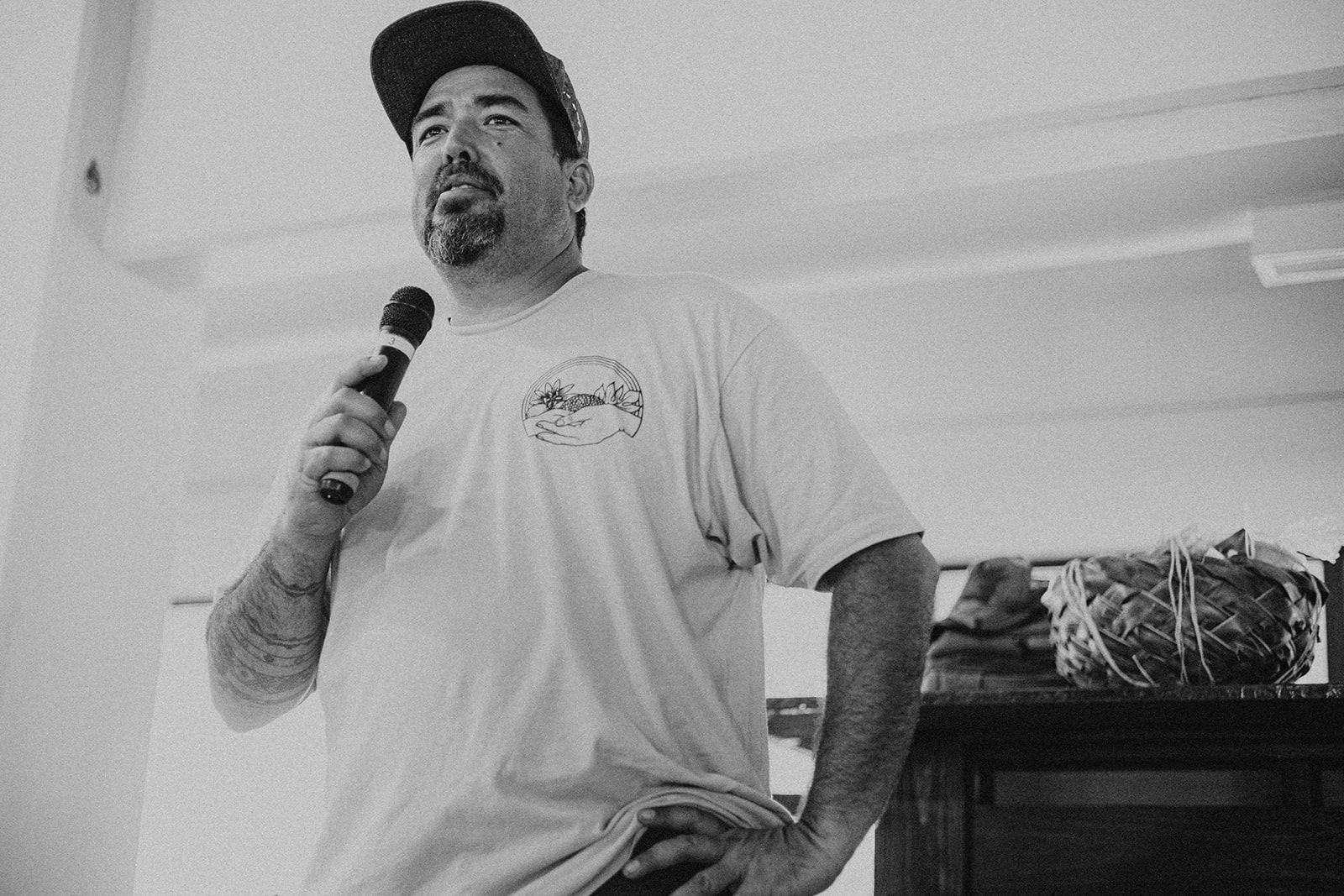 Man speaking into a microphone indoors, wearing a cap and T-shirt.