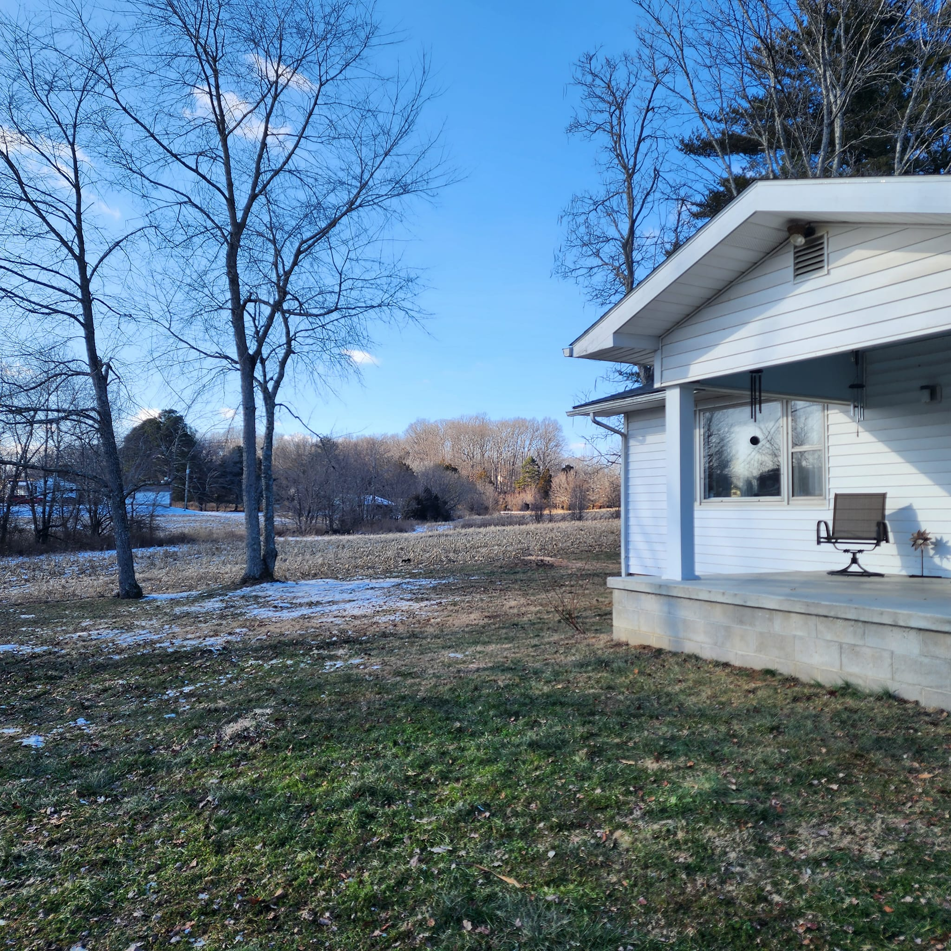A white house with a porch on a grassy field with trees and a blue sky.