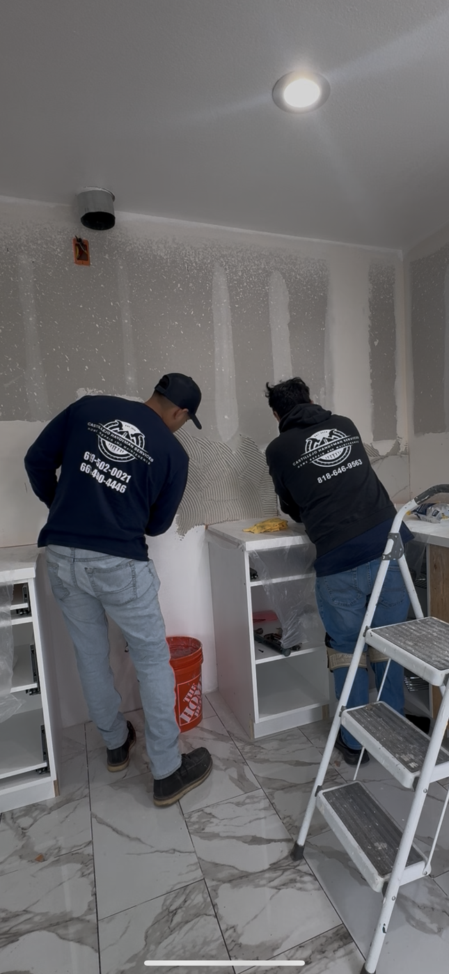 Two workers tiling a kitchen, wearing blue shirts, cabinets, ladder, red bucket, tile floor.