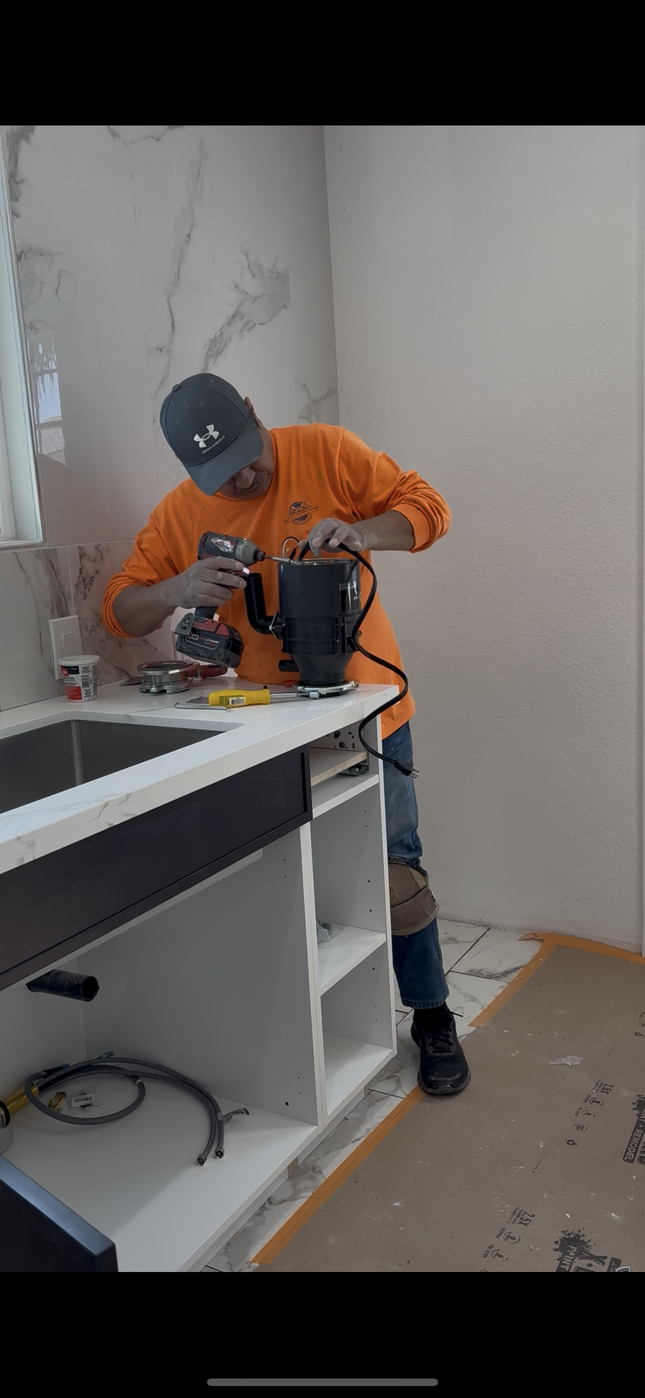 A person in an orange shirt uses a tool on a countertop near a sink, inside a room.