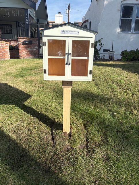 Small, white library box with brown doors on a wooden post in a grassy yard.
