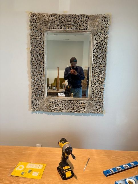 A man taking a selfie in a decorative framed mirror hanging on a white wall; tools on a table below.