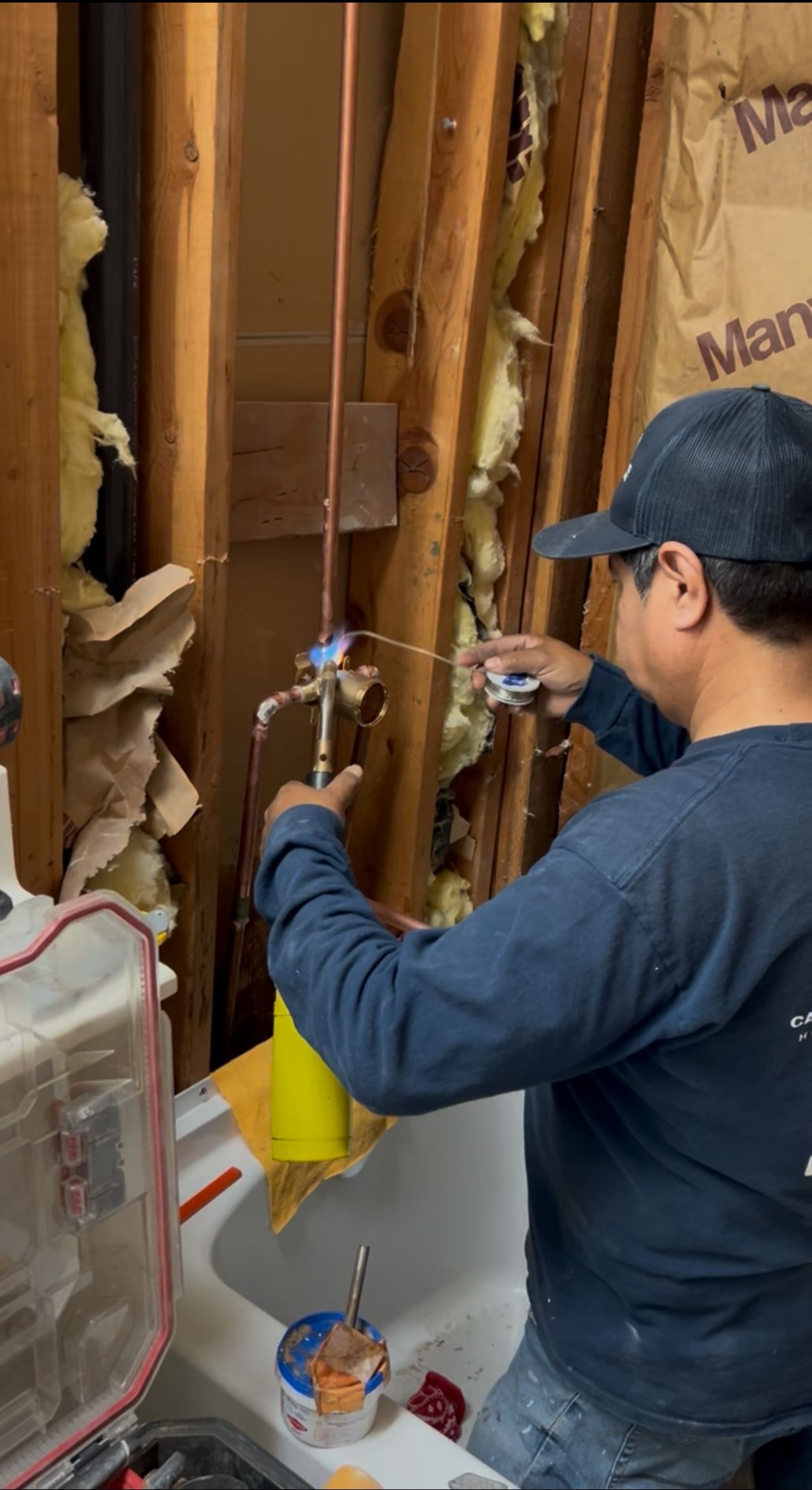 A person in a blue shirt and hat working on copper plumbing pipes within a wall.