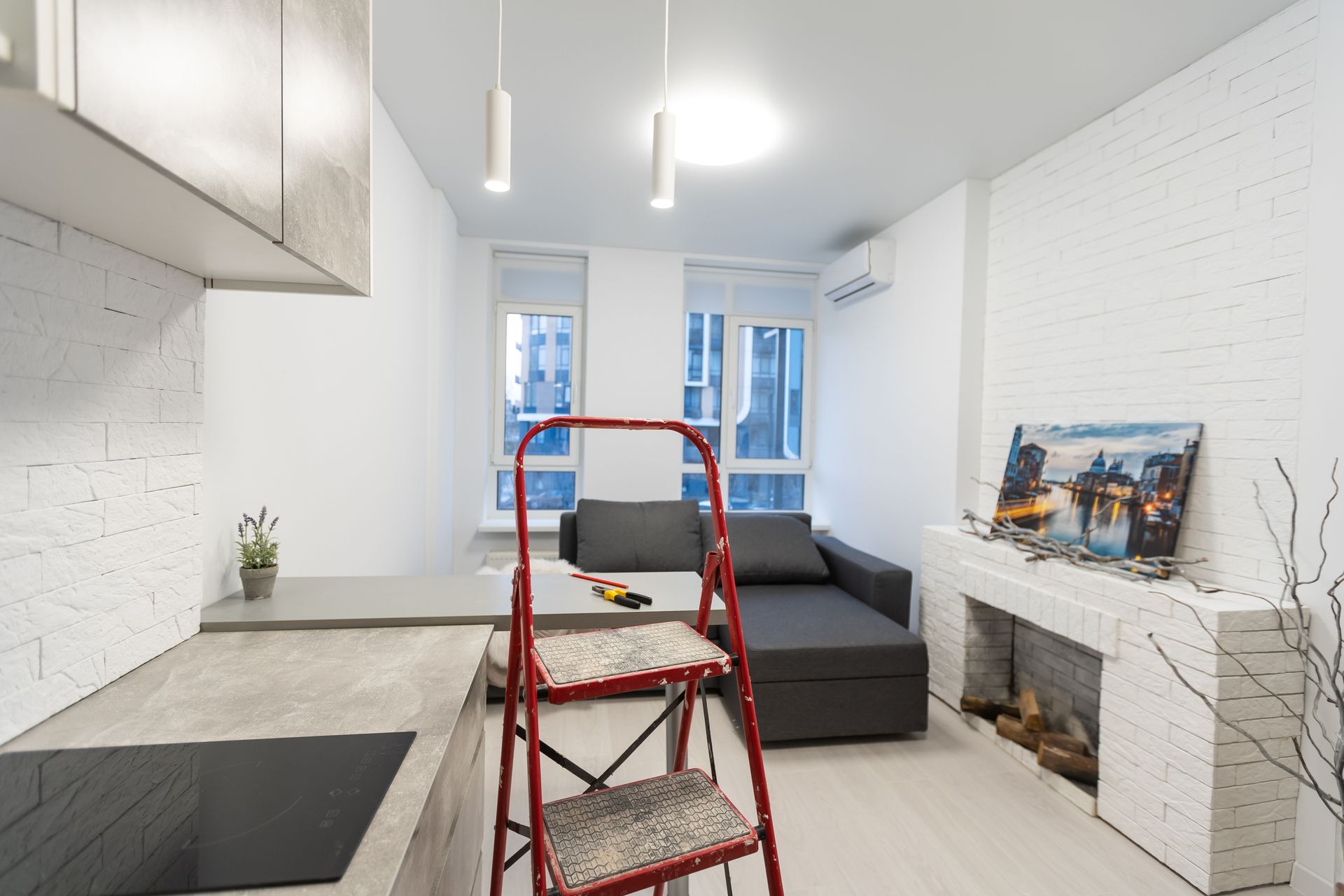 Bright white studio with a red step ladder, kitchen counter, gray sofa, and fireplace.