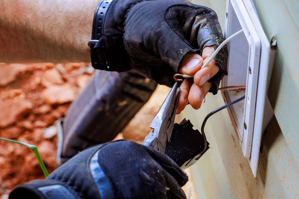 Gloved hands using a cable tester on outdoor wall wiring near an electrical box