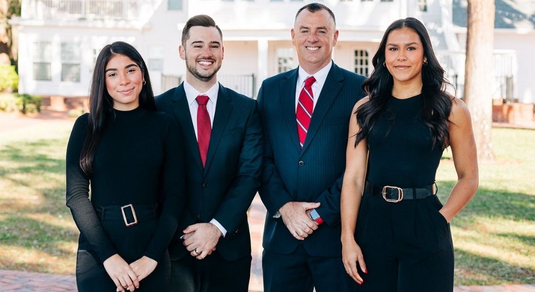 Four people stand outdoors in a row, wearing professional business attire, posing for a group portrait in front of a house.