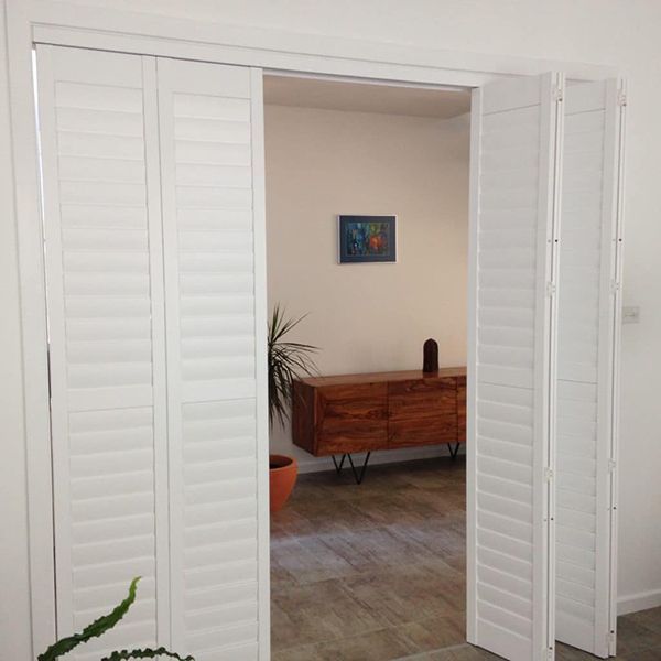 White Folding Shutters Opening Into A Room With A Cabinet And Plant — Winning Blinds in Cardiff, NSW