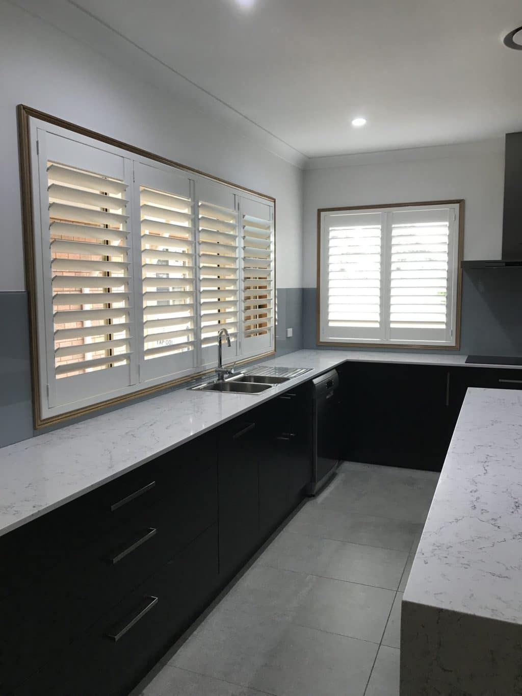 A kitchen with black cabinets and white counter tops and shutters on the windows — Winning Blinds in Cardiff, NSW