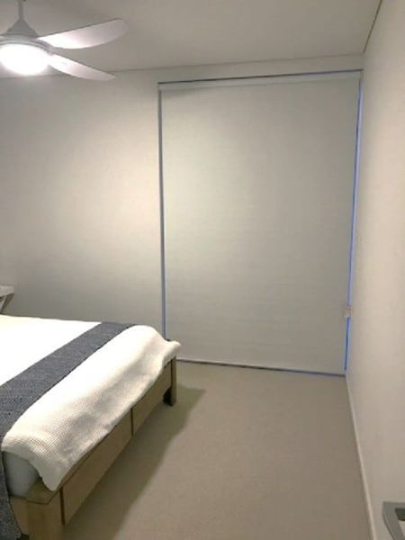 Bedroom With A White Roller Blind Covering A Large Window — Winning Blinds in Cardiff, NSW