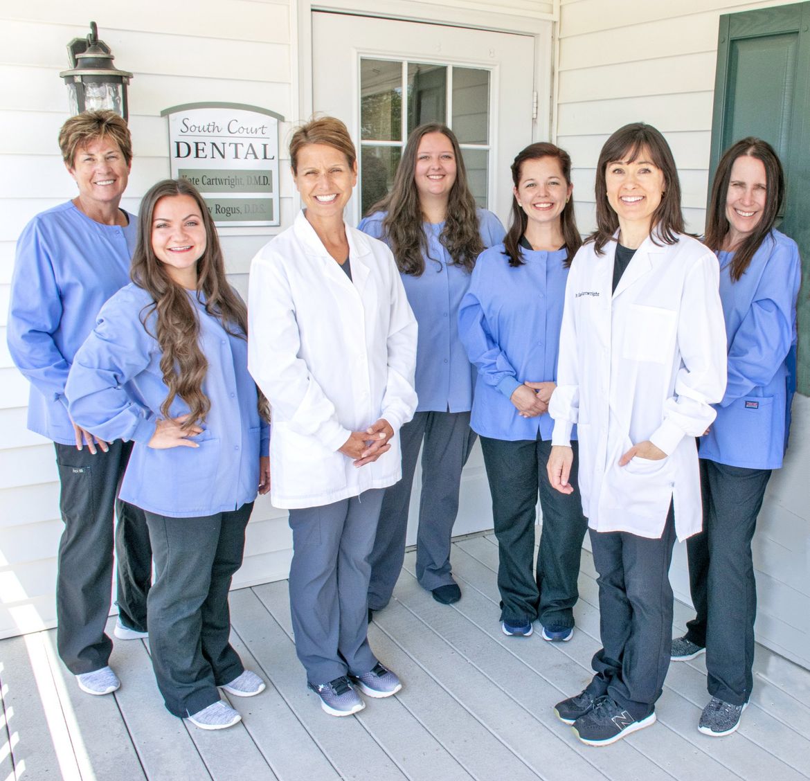 Dental office staff standing on a porch. They wear scrubs and lab coats. Smiles are visible.