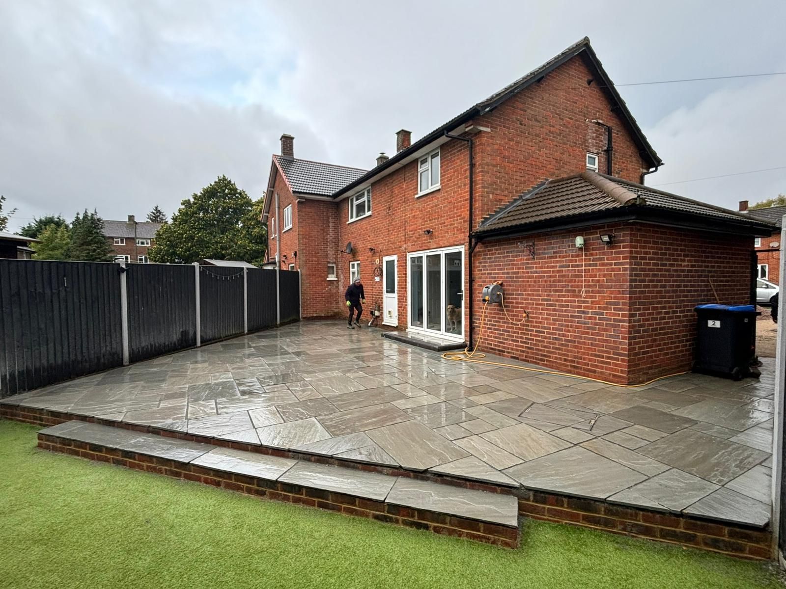 Backyard patio with brick house, fence, and artificial grass; wet ground.