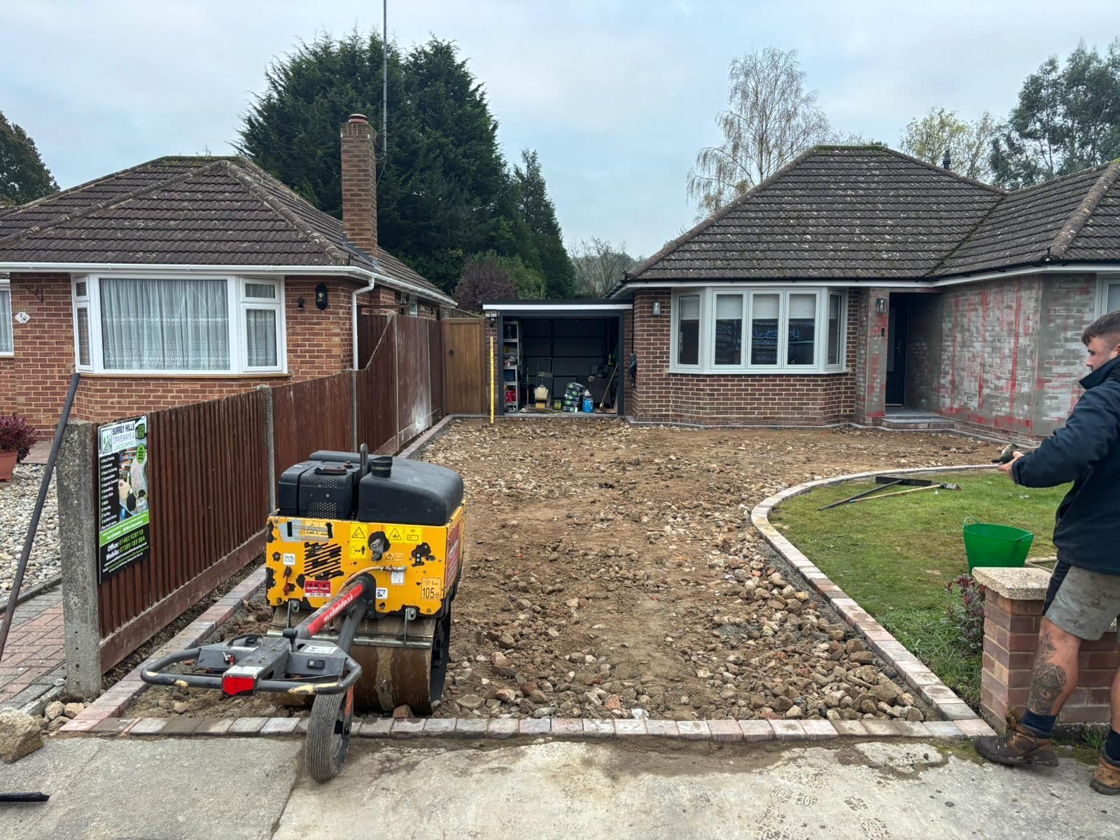 Driveway excavation and site preparation in Woking by Surrey Hills Driveways & Landscaping, showing a worker using a compactor between two houses.