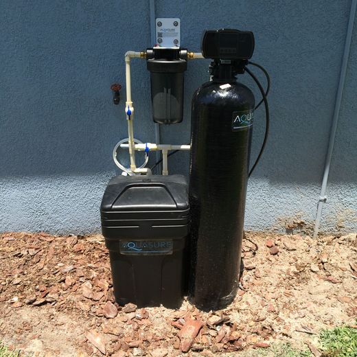 A residential water filtration system with a tall black tank, a smaller black brine tank, and a filter, set on dirt.