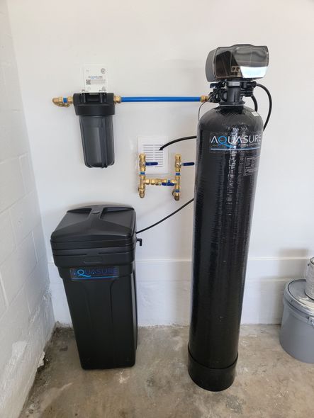 Water softener system with a tall black tank, a small brine bin, a pre-filter, and brass plumbing pipes against a wall.