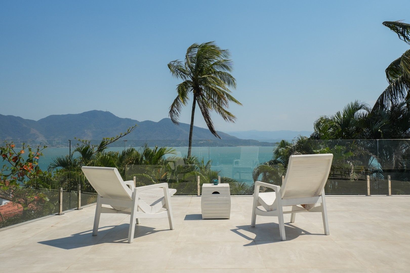 Duas espreguiçadeiras brancas de frente para um oceano tropical com uma palmeira e montanhas sob um céu azul límpido.
