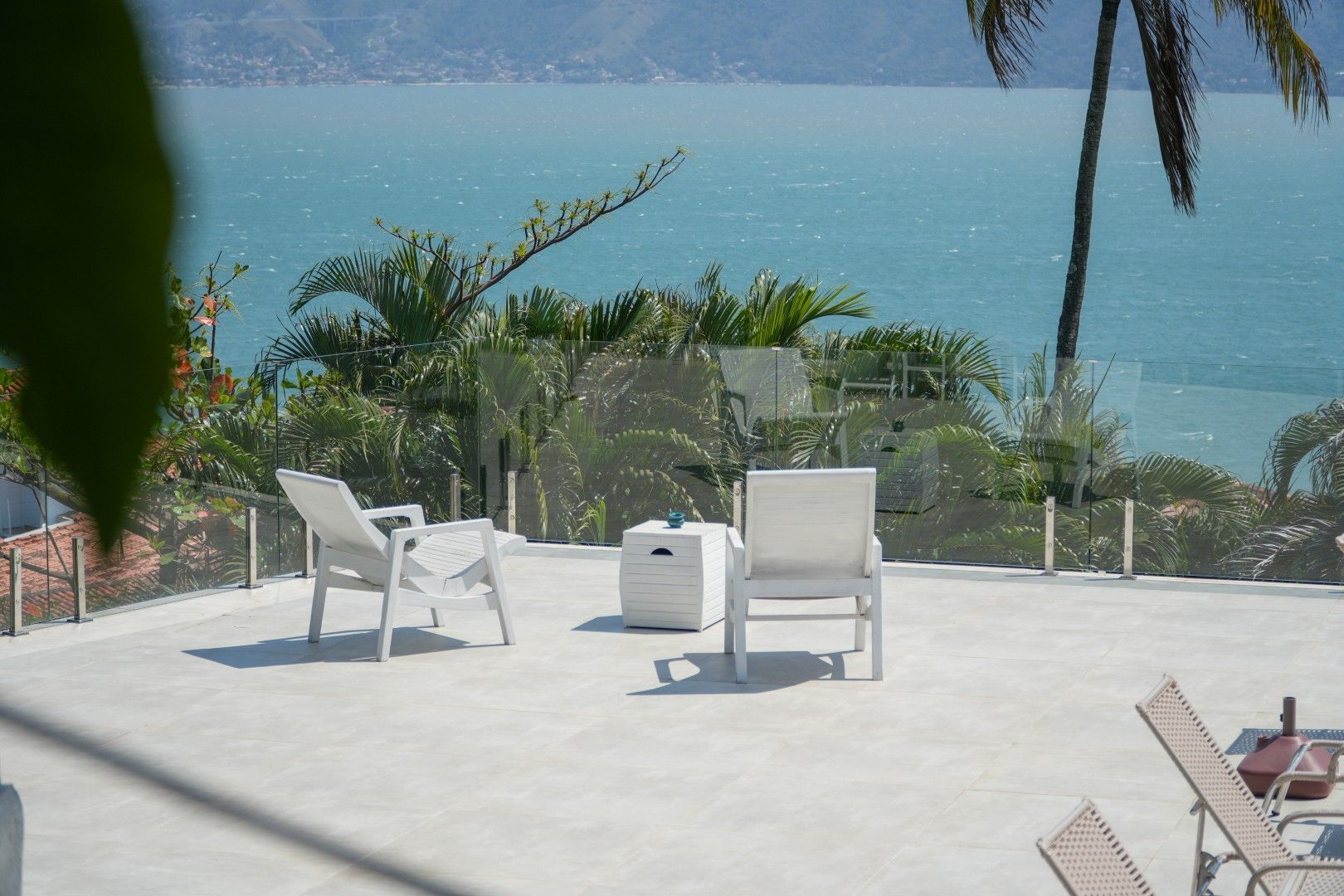 Cadeiras de jardim brancas e uma pequena mesa em um terraço ensolarado com vista para a água azul e colinas ao longe.