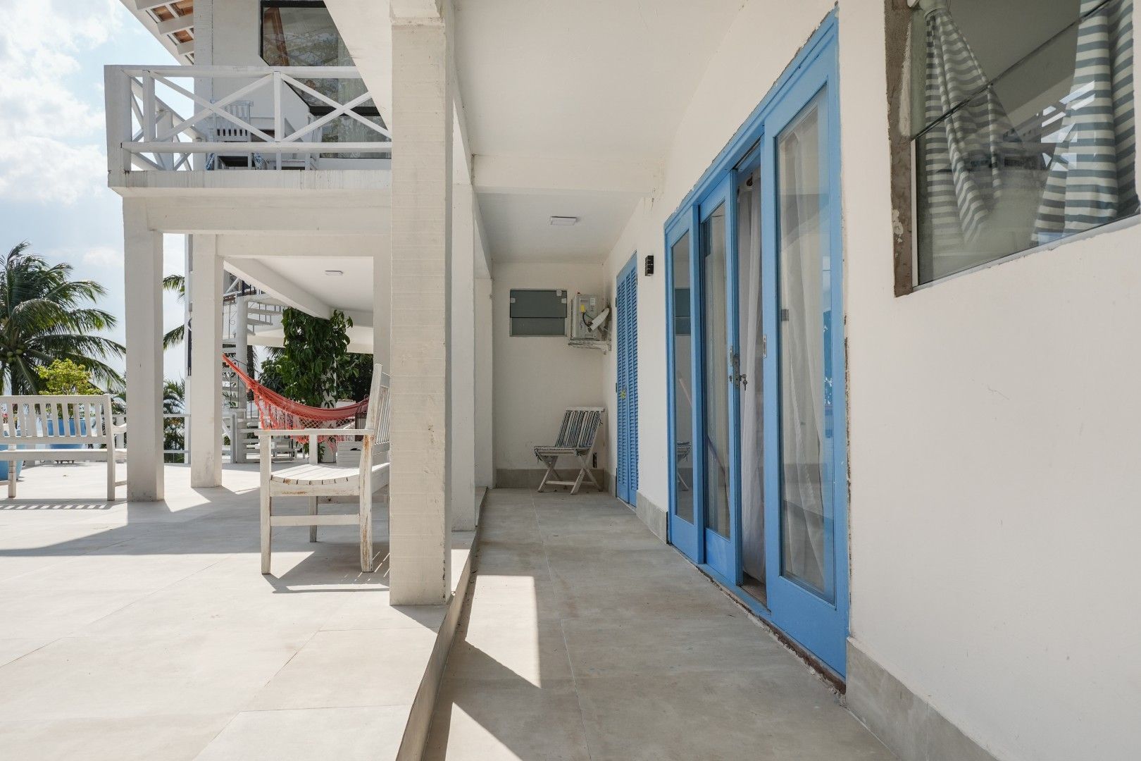 Pátio branco com portas e janelas azuis, colunas brancas e uma rede em um ambiente tropical luminoso.