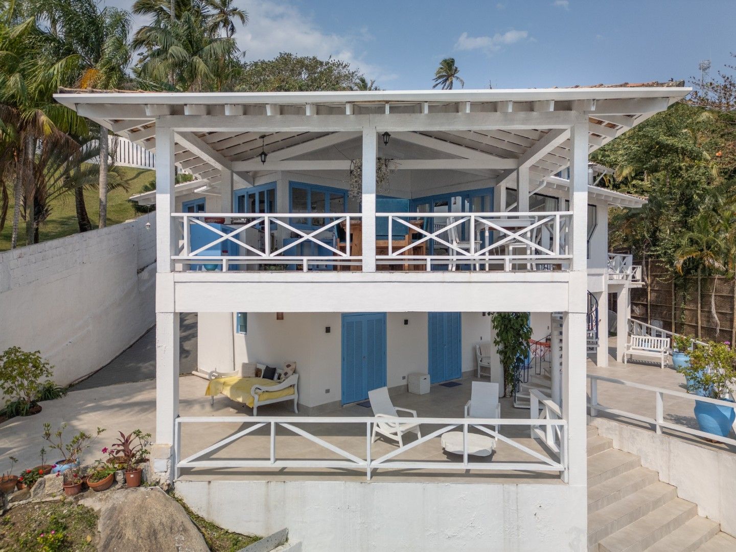 Casa de praia branca de dois andares com portas azuis, varandas e terraço com vista para o mar ao lado de palmeiras.