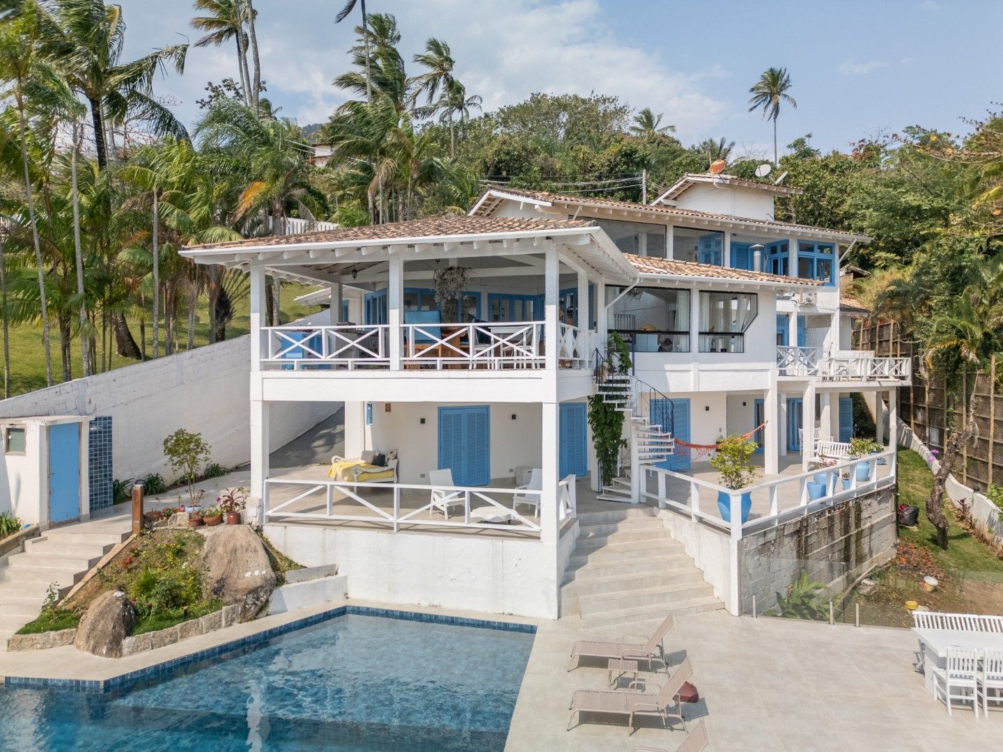 Vila tropical branca com detalhes em azul e piscina em uma encosta, rodeada por palmeiras.