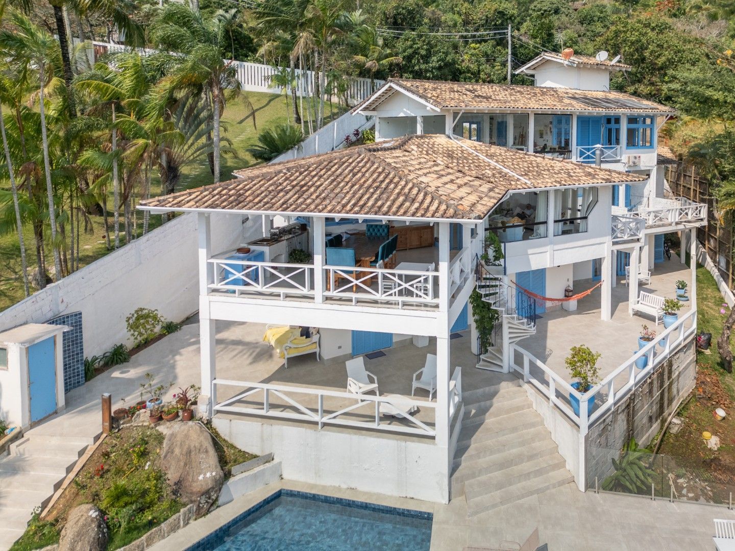Casa branca à beira-mar com detalhes em azul, telhado de telha e varandas que circundam a fachada, situada em uma encosta.