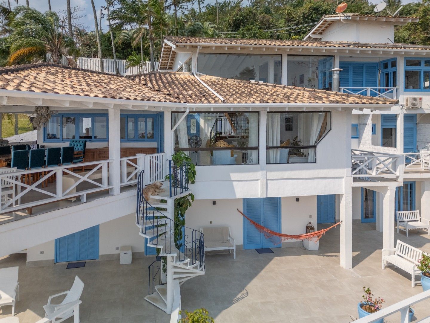 Moradia branca à beira-mar com persianas azuis, telhados de telha e varandas com vista para um pátio de areia.