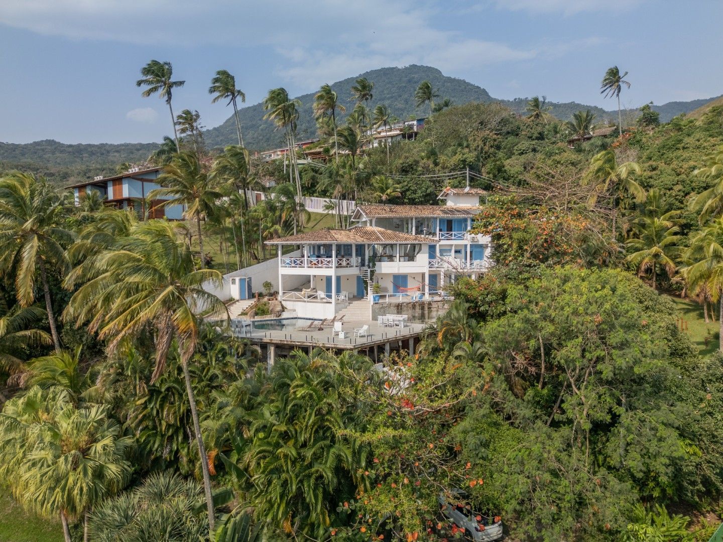 Vila branca na encosta de uma colina, em meio à exuberante vegetação tropical, com palmeiras e montanhas ao longe.