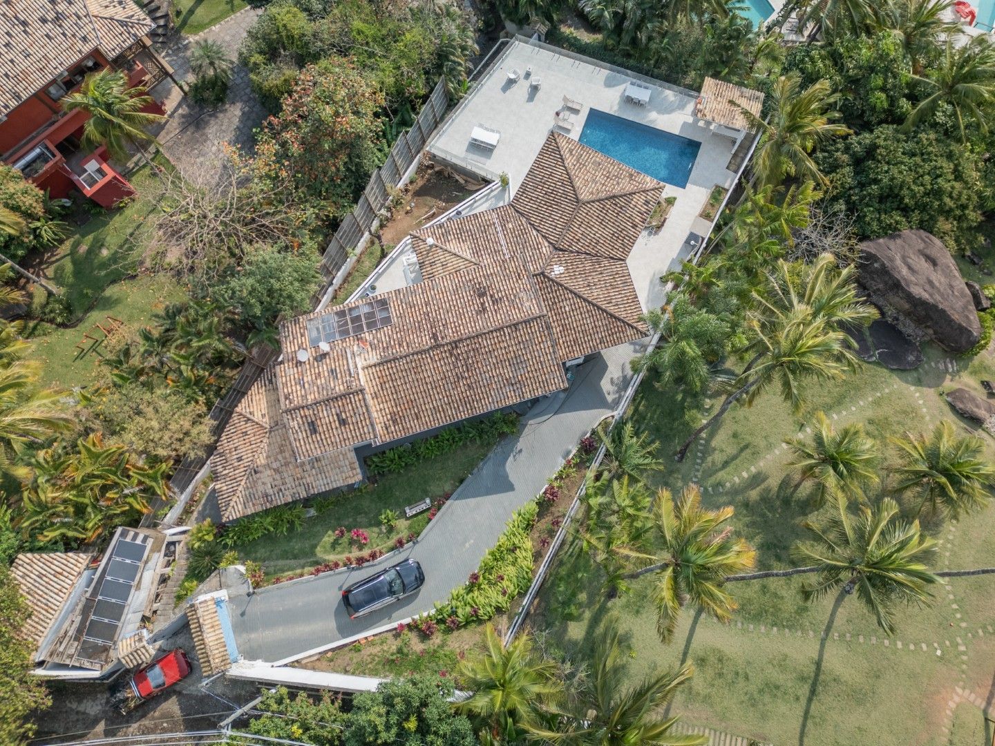 Vista aérea de uma casa tropical com telhado de telha, entrada para carros, piscina e palmeiras.