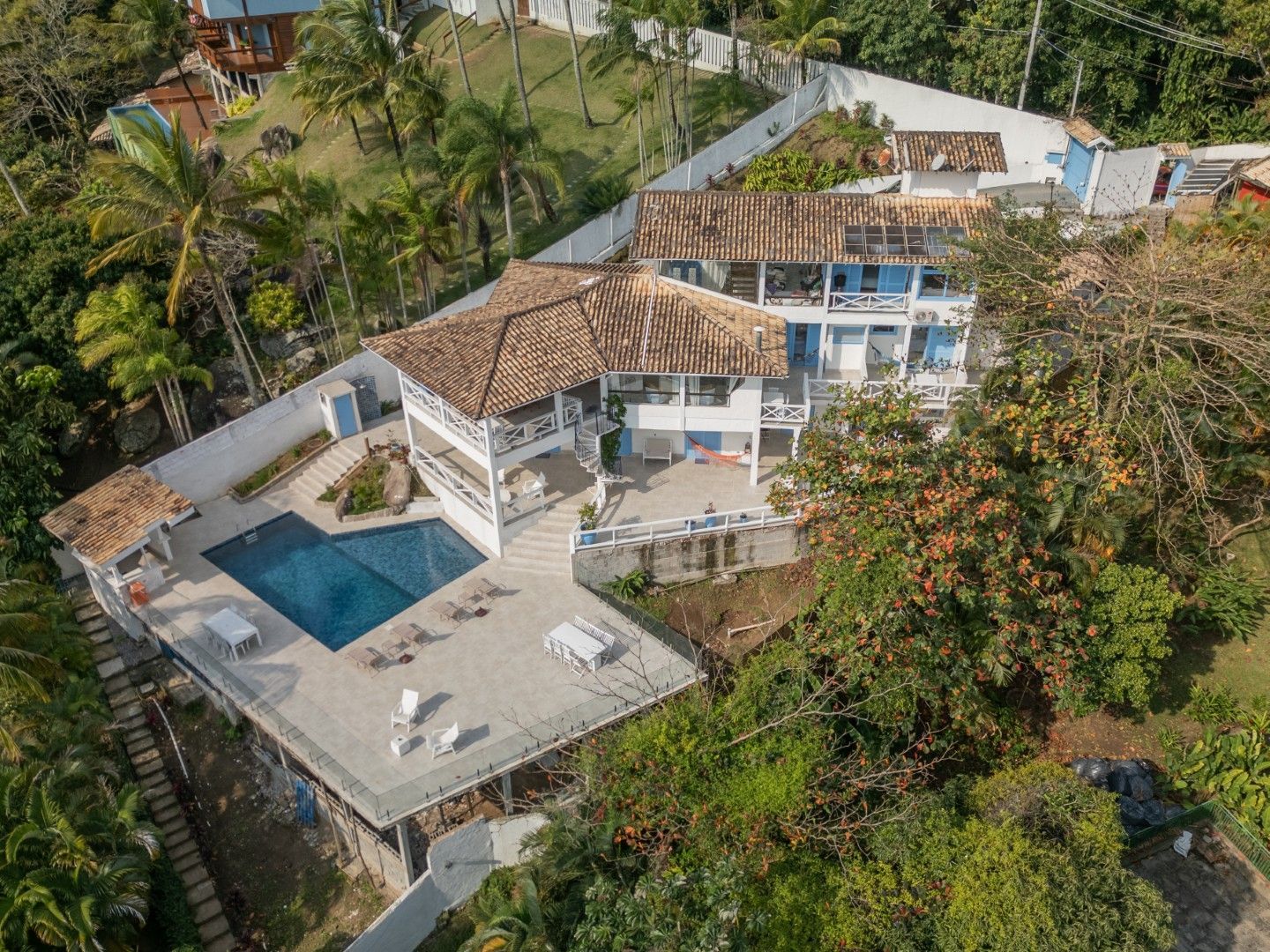 Vista aérea de uma casa branca com telhados de telha vermelha, piscina azul e terraço rodeado por vegetação tropical.