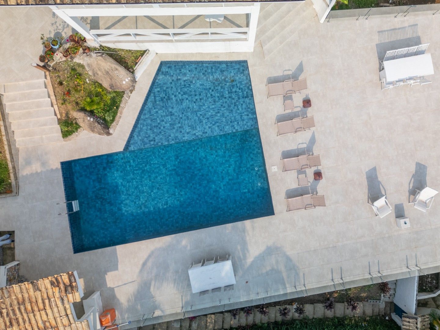 Vista aérea de uma piscina com espreguiçadeiras em um pátio ensolarado ao lado de um jardim.
