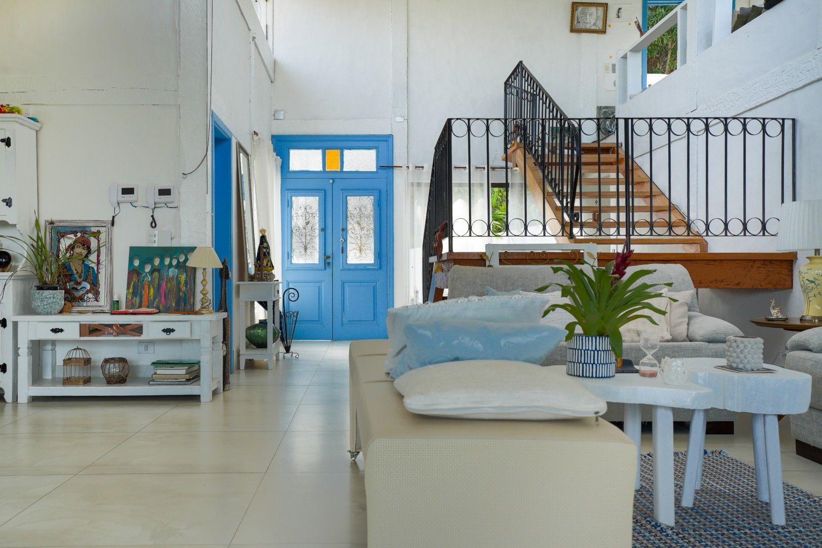Sala de estar iluminada com porta de entrada azul, sofá branco, escada e plantas.