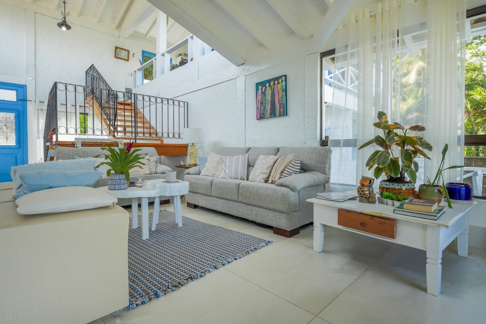 Sala de estar ampla e iluminada com vigas brancas, sofá cinza, detalhes em azul e plantas de interior.