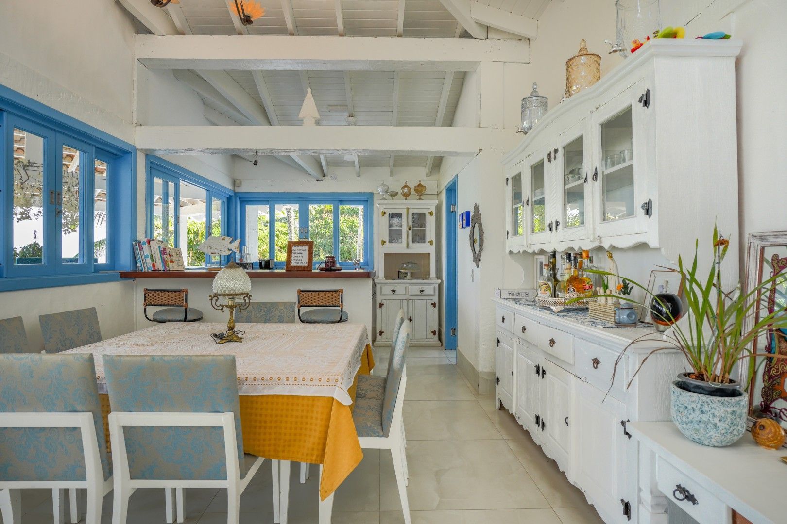 Sala de jantar e cozinha totalmente brancas e iluminadas, com janelas azuis, armários brancos e uma mesa posta com toalha de mesa amarela.